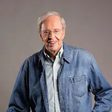 A man in blue shirt and jeans standing next to wall.